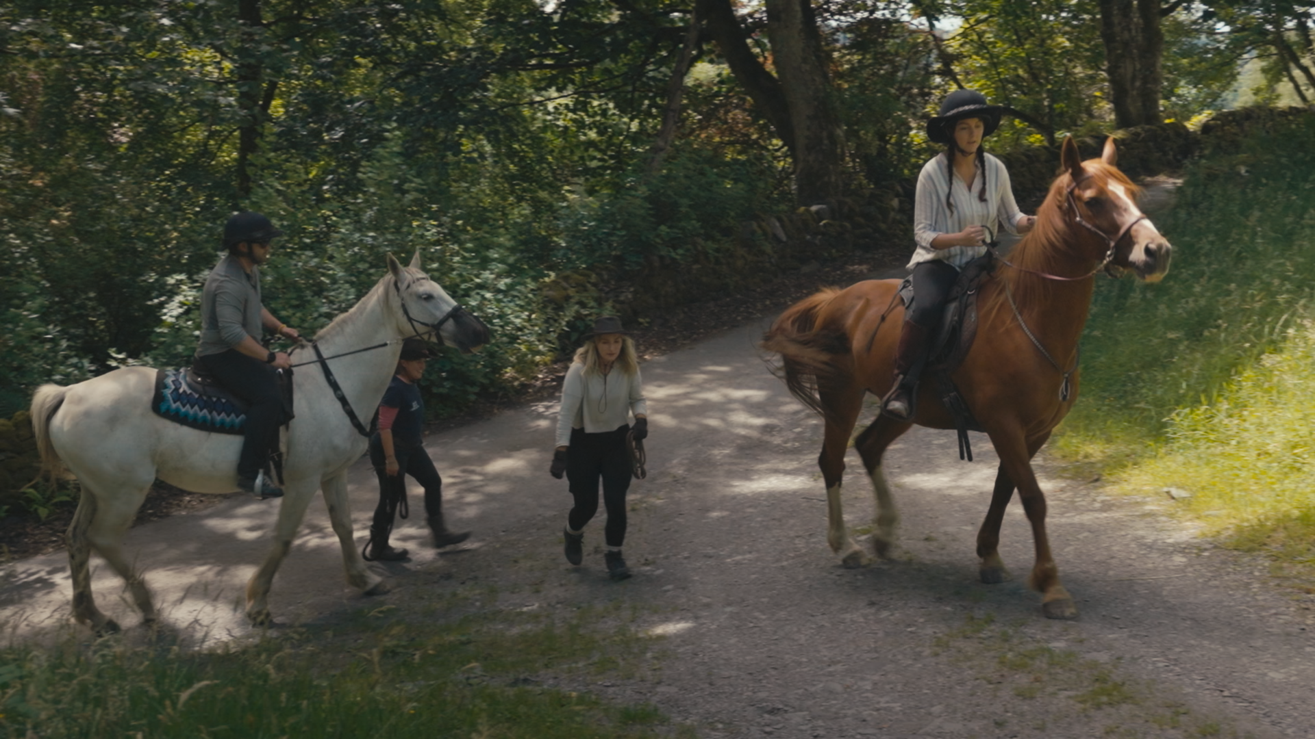 Two riders on their horses clop along a country path while a couple of people walk alongside