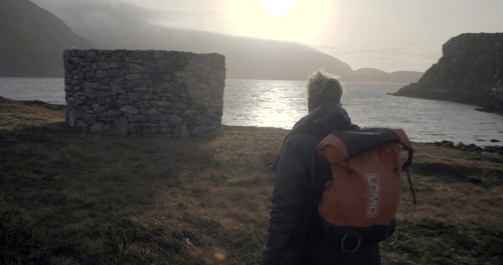 A person wearing a backpack faces a stone wall near the sea