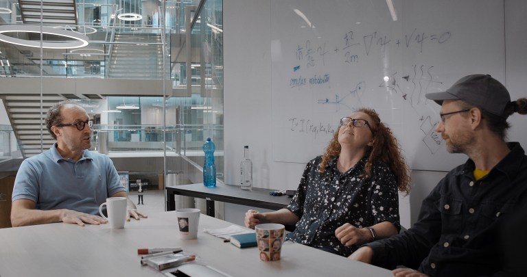 Three people sit chatting around a desk in a modern looking office, whiteboard behind with formulae jotted on it