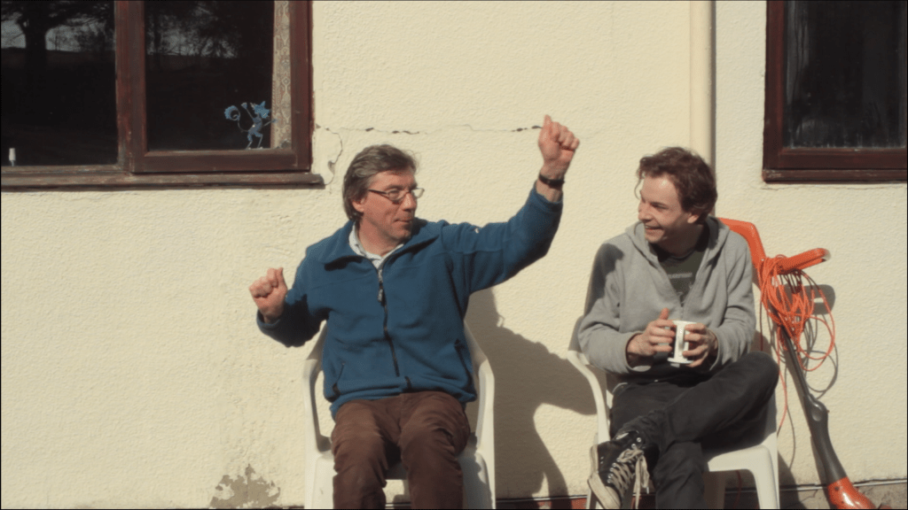Two men have a relaxed looking chat in the sun at the back of a house, sitting in garden chairs.