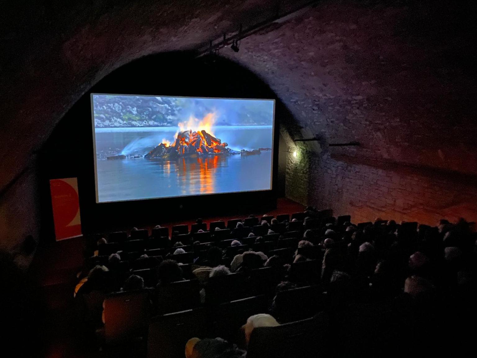 An audience watches the film in what appears to be a large railway arch space or something similar